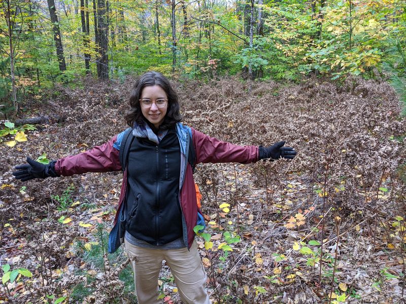 Photo of a white nonbinary person extending their arms to the side, gesturing to the forest