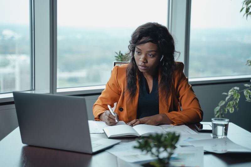 Photo of a Black person looking at a laptop and writing notes in a very nice office with windows behind them