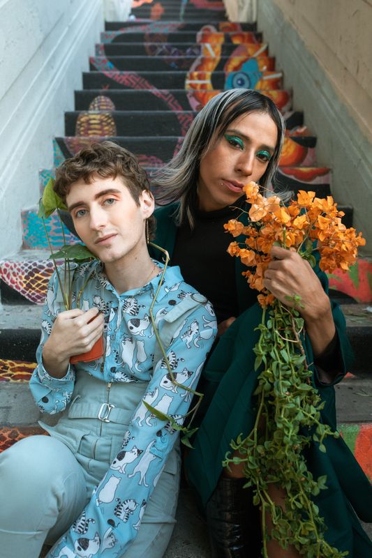 Two people with pale and medium skin tone, wearing eyeshadow and holding plants and flowers, sit on a staircase