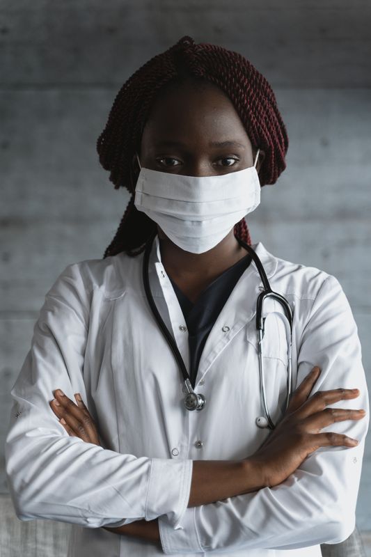 Dark-skinned person with braided hair wearing a cloth face mask and white medical coat with stethoscope