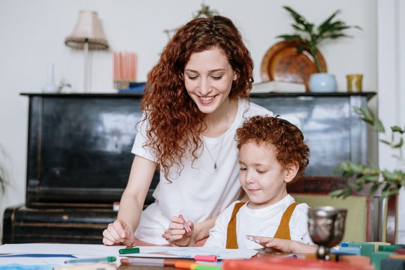 A curly-haired adult sits with a curly-haired child, smiling while drawing a picture.