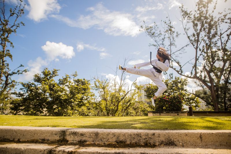 Person wearing a martial arts gi, jumping while kicking to the side, outside on a sunny day