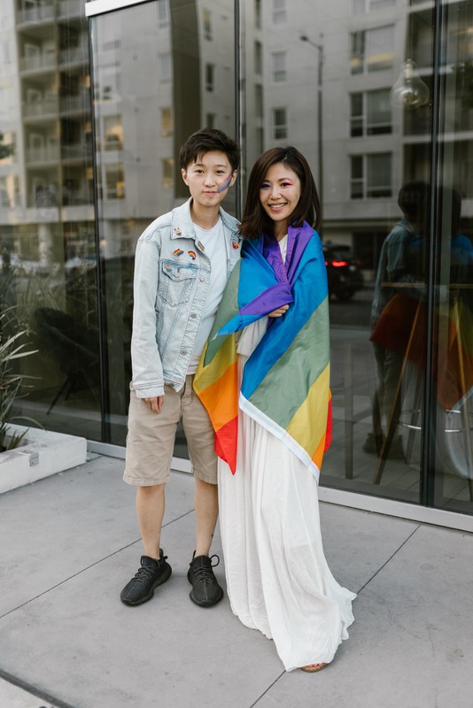 Photo of couple standing together. One person is wearing a rainbow flag around their shoulders.