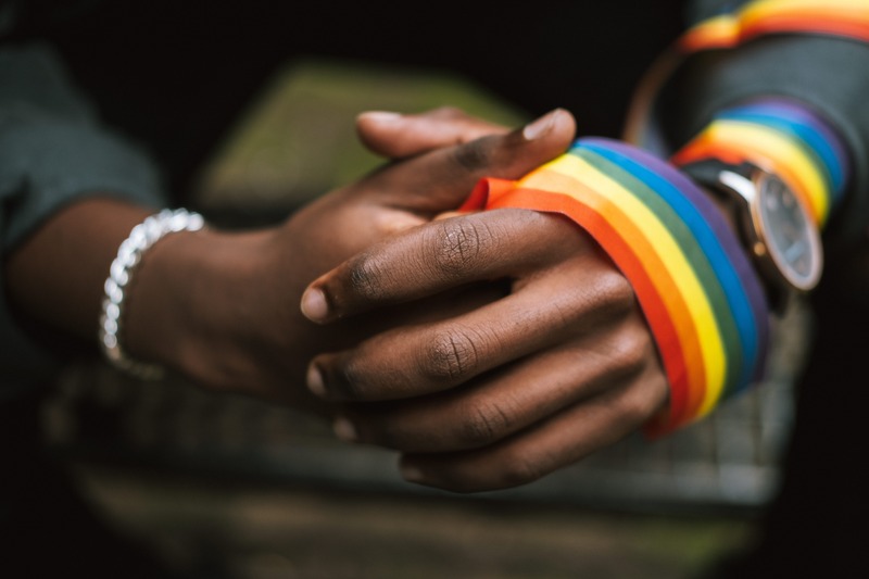 Photo of a dark skinned person, close up on their hands clasped together, with a rainbow ribbon wrapped around one hand and wrist