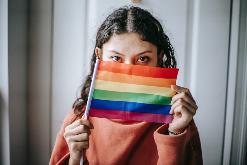 Person with light skin and black curly hair holding a small rainbow flag in front of them